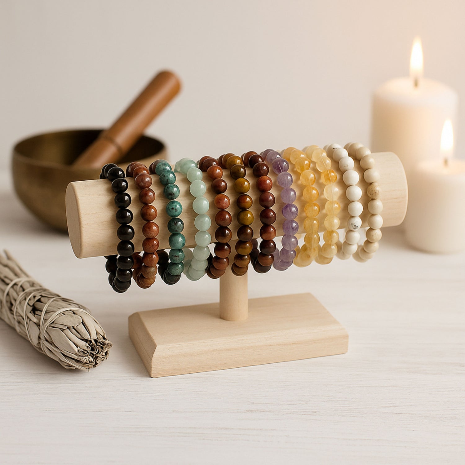 Présentoir élégant avec plusieurs bracelets de lithothérapie en pierres naturelles, disposé dans une ambiance ésotérique apaisante. On distingue de la sauge, un bol chantant tibétain et des bougies, créant un décor spirituel et raffiné.