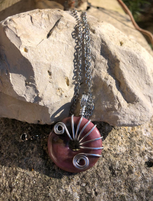 Pendentif spirituel artisanal en Rhodonite et métal posé sur une roche claire