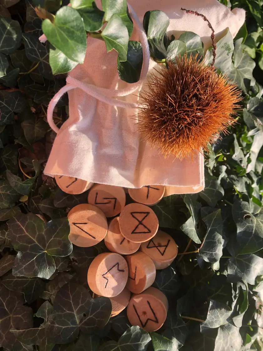 Runes en bois de cerisier pyrogravées sortant d'une pochette en coton, posées sur du lierre